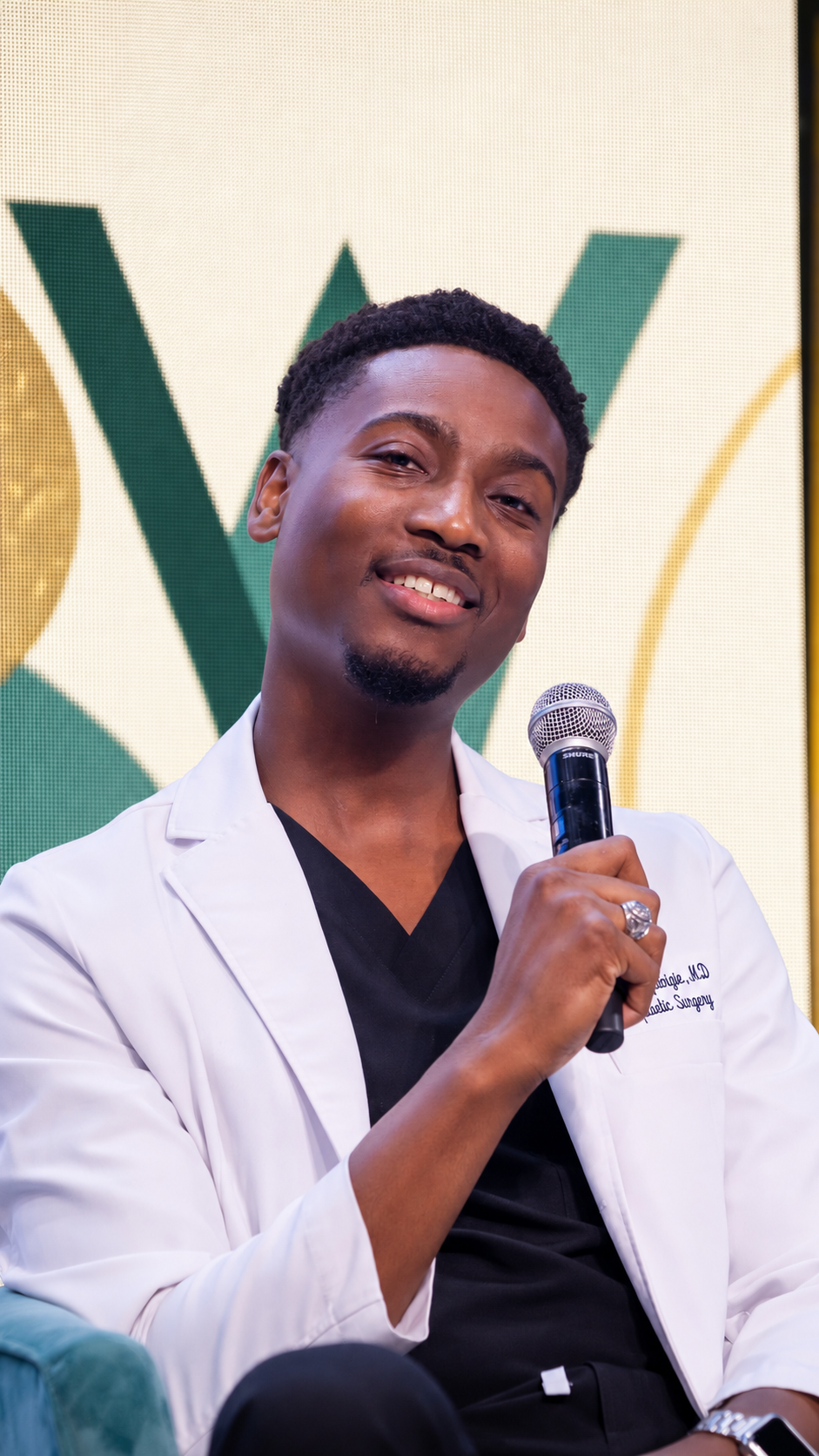 Dr. Iggy in a white coat speaking into a microphone before a green-and-gold stage backdrop
