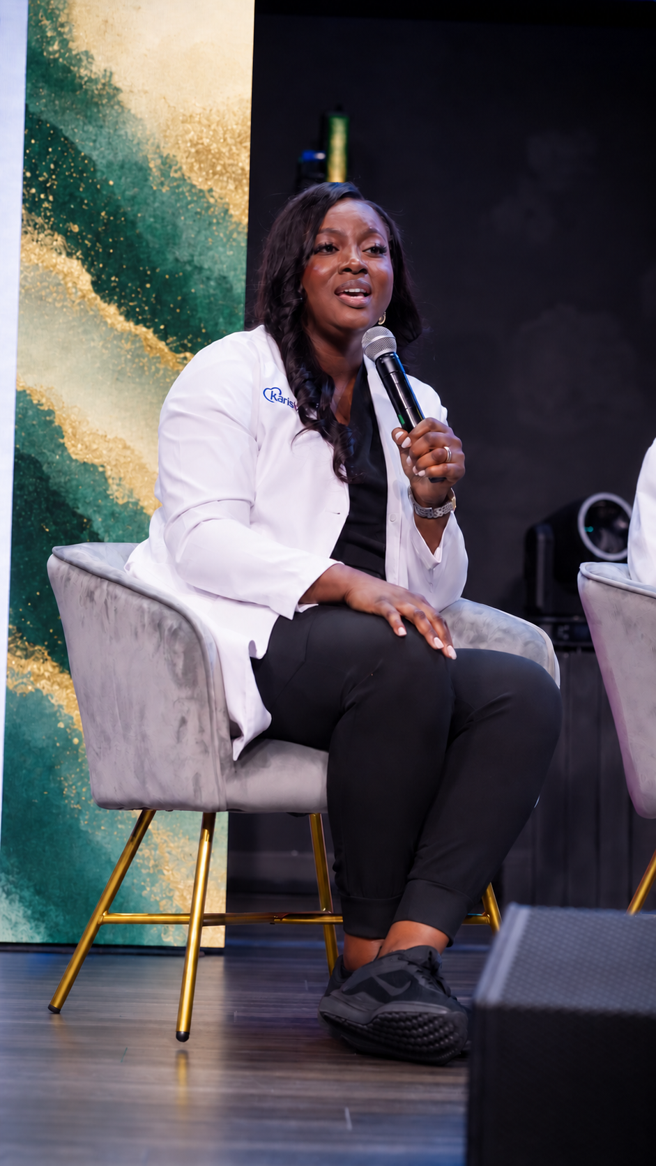 Dr. Vera seated on stage in a white coat, speaking into a microphone