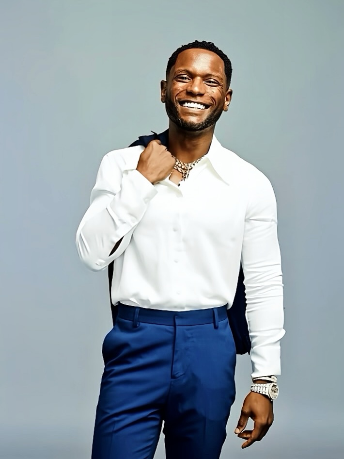 Dr. Rose smiling in a white open-collar shirt and blue trousers, with a dark jacket over his shoulder against a pale blue-gray background